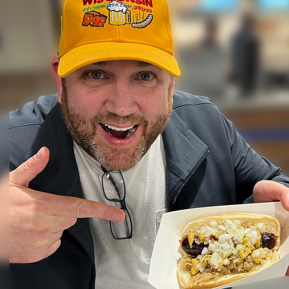 Image of Joe wearing a bright yellow baseball cap, pointing at a bratwurst sandwich with mustard and onions. 