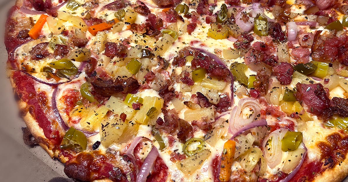 Partial image of Lake Country Supreme thin-crust pizza topped with giardiniera, pineapple, bacon, and onion, sitting in a pizza box.