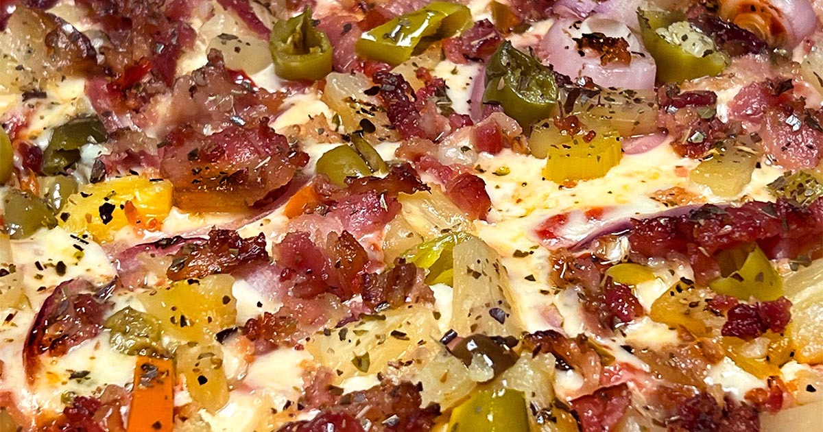 Closeup image of Lake Country Supreme thin-crust pizza taken from the side shows crust, sauce, giardiniera, pineapple, bacon, and onion